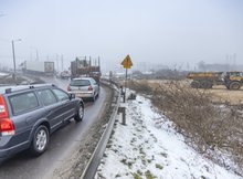 Po lewej stronie auta stojące przed zamkniętym przejazdem kolejowym, po prawej maszyny na budowa wiaduktu. 